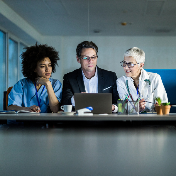 Our team draws upon decades of industry experience and works closely with our clients A female in blue scrubs, a man in a business suit on a laptop, and a woman in a physician's coat all looking at the business man's laptop screen.